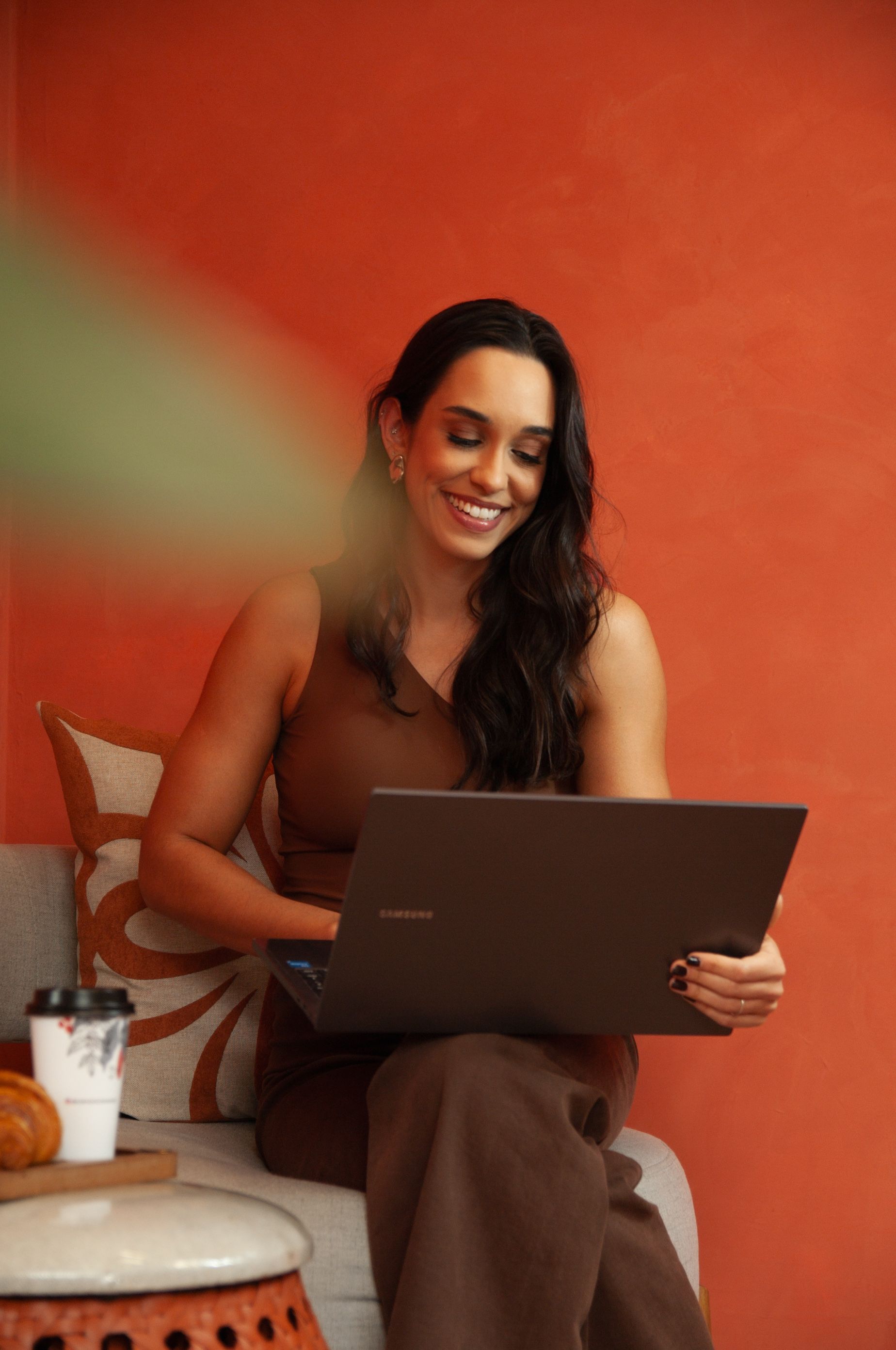 Bia sorrindo enquanto trabalha no notebook, com um café e pão na mesa lateral, contra fundo terracota